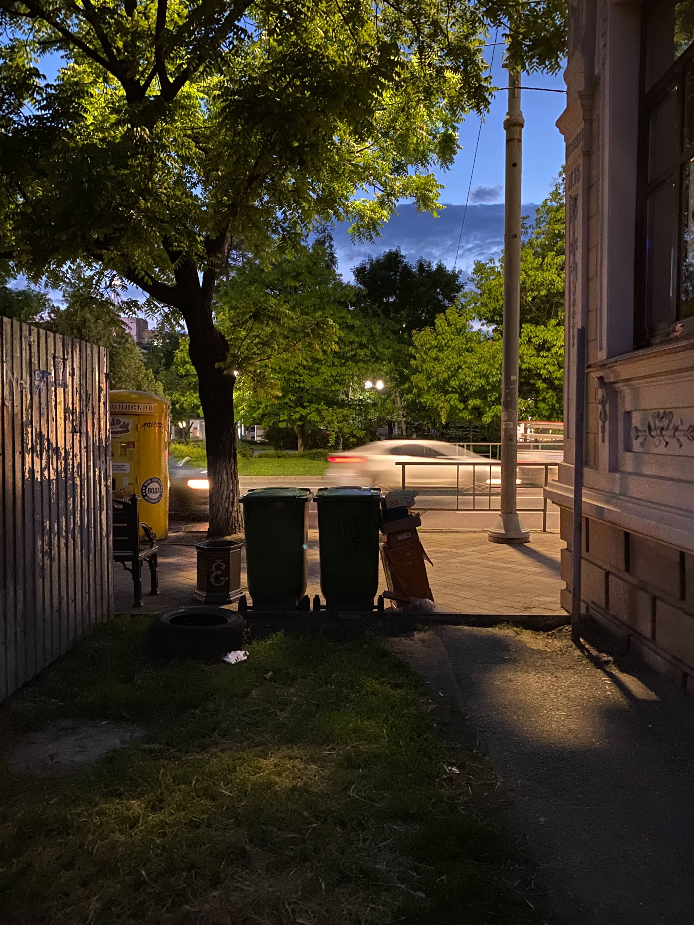 Вид из тёмного проулка на оживлённую улицу. Посередине кадра силуэты урн
